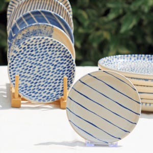 May include: A selection of ceramic plates with blue and white patterns. The plates showcase diverse designs, such as stripes and dotted motifs. The plates are presented on a wooden rack and a clear plastic stand, highlighting their decorative nature.
