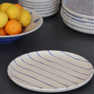 May include: White oval serving platter with blue diagonal stripes. The ceramic dish is part of a set, with other plates and bowls visible. A bowl of lemons and oranges is in the background. The tableware has a handmade aesthetic.