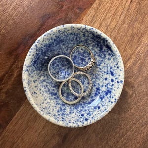 May include: A small white ceramic bowl with a blue speckled pattern, containing several silver rings.