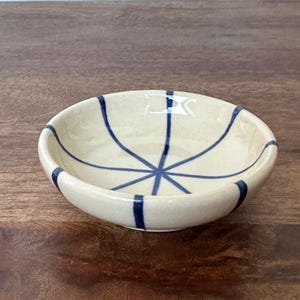 May include: A small, white ceramic bowl with a blue and white striped design. The bowl is round and has a slight lip around the edge.