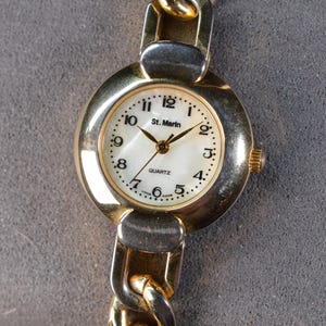 Puede incluir: Un reloj de cuarzo St. Marin dorado con esfera blanca y agujas doradas. El reloj tiene una correa de eslabones dorados y una esfera redonda con números negros. La esfera del reloj dice "St. Marin" y "Quartz".