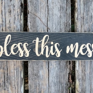 May include: A black wooden sign with the words "bless this mess" written in gold paint.
