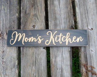 May include: A black wooden sign with the words "Mom's Kitchen" written in gold cursive lettering.