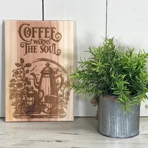 May include: Wooden sign with the text "Coffee Warms The Soul" and a coffee pot illustration. A small green plant in a galvanized metal pot sits to the right of the sign. The sign and plant are on a white wooden surface.