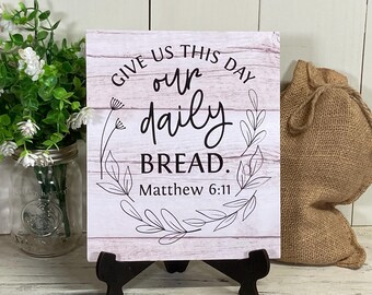 May include: White wooden sign with black text that reads "Give us this day our daily bread. Matthew 6:11". The sign is decorated with black floral designs and is sitting on a small black easel.
