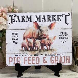 May include: A white wooden sign with the text "FARM MARKET" in black lettering. The sign features a painted image of three pink pigs and the text "OPEN 9 to 5 DAILY", "FRESH ORGANIC PRODUCE", "MEATS EGGS HONEY CHEESE DAIRY", and "PIG FEED & GRAIN".