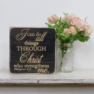 May include: A black wooden sign with a gold inscription that reads "I can do all things through Christ who strengthens me. Philippians 4:13". The sign is sitting on a white shelf with a glass jar of pink flowers.