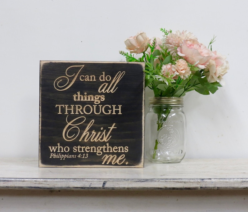 May include: A black wooden sign with a gold inscription that reads "I can do all things through Christ who strengthens me. Philippians 4:13". The sign is sitting on a white shelf with a glass jar of pink flowers.