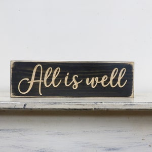 Puede incluir: Un letrero de madera negro con las palabras "All is well" en una fuente dorada.