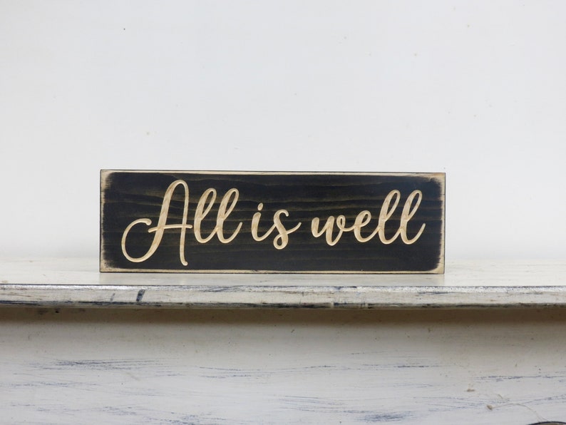 Puede incluir: Un letrero de madera negro con las palabras "All is well" en una fuente dorada.