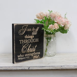 May include: A black wooden sign with a white inscription that reads "I can do all things through Christ who strengthens me. Philippians 4:13". The sign is sitting on a white wooden surface with a glass jar of pink flowers.