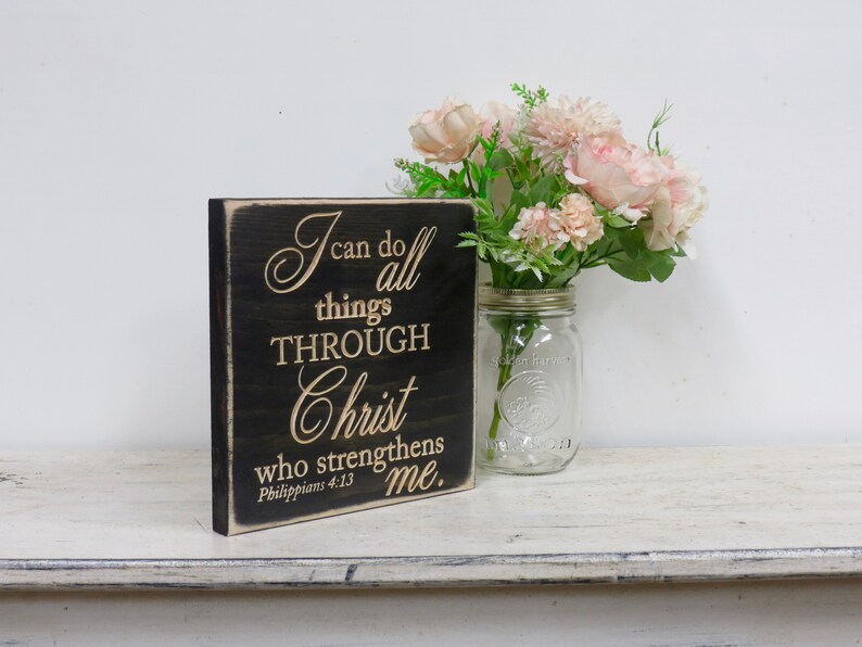 May include: A black wooden sign with a white inscription that reads "I can do all things through Christ who strengthens me. Philippians 4:13". The sign is sitting on a white wooden surface with a glass jar of pink flowers.