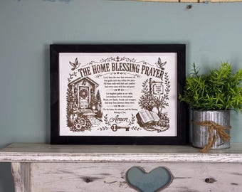May include: A rectangular black frame containing a white print with the text THE HOME BLESSING PRAYER. The design features a house entrance, a bird, a key, an open book, and floral accents in dark brown ink on a light background.