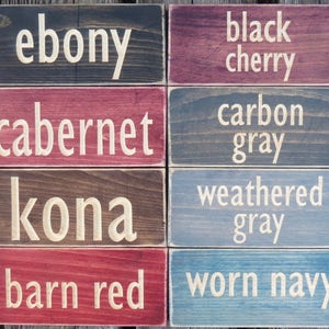 May include: Eight wooden signs with different wood stains and paint colors. The signs have the following text: ebony, black cherry, cabernet, carbon gray, kona, weathered gray, barn red, worn navy.