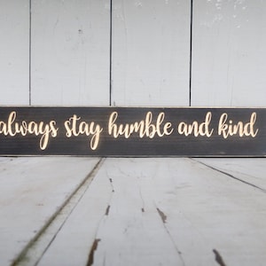 May include: Black wooden sign with the words "always stay humble and kind" written in gold lettering.