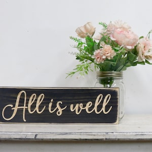 Puede incluir: Un letrero de madera negro con las palabras "All is well" escritas en una fuente dorada. El letrero est&aacute; sentado en un estante blanco con un jarr&oacute;n de flores rosas en el fondo.