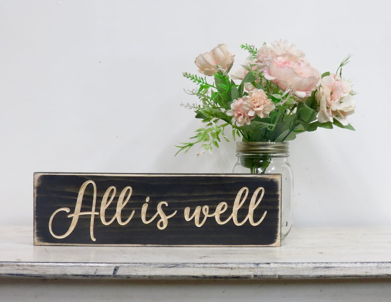 Puede incluir: Un letrero de madera negro con las palabras "All is well" escritas en una fuente dorada. El letrero est&aacute; sentado en un estante blanco con un jarr&oacute;n de flores rosas en el fondo.
