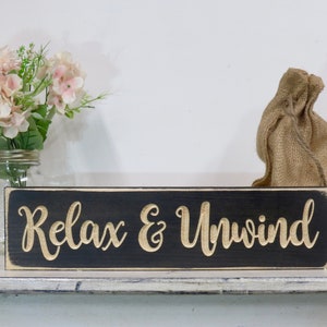 May include: Black wooden sign with white trim and the words "Relax & Unwind" in gold lettering.