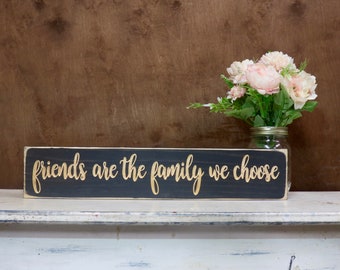 May include: Black wooden sign with white distressed paint and gold lettering that reads "Friends are the family we choose".