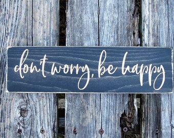 May include: A dark wood sign with the text "don't worry, be happy" in white cursive lettering.
