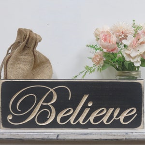 May include: A black wooden sign with white lettering that reads "Believe". The sign is sitting on a white shelf with a burlap bag and a bouquet of pink flowers in a glass jar behind it.
