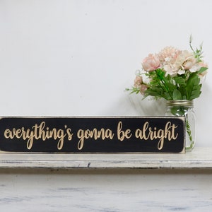 May include: Black wooden sign with the text "everything's gonna be alright" in gold lettering. The sign is sitting on a white shelf with a vase of pink flowers in the background.