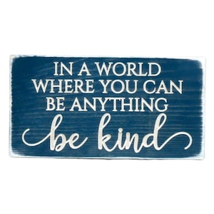 May include: A blue wooden sign with white lettering that reads "In a world where you can be anything be kind".
