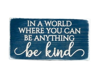 May include: A blue wooden sign with white lettering that reads "In a world where you can be anything be kind".