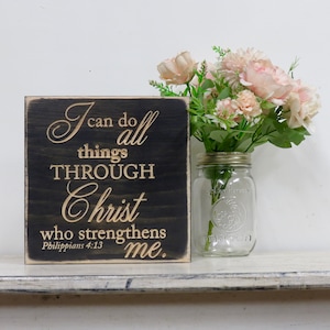 May include: A black wooden sign with the text "I can do all things THROUGH Christ who strengthens me. Philippians 4:13" in gold lettering. The sign is sitting on a white shelf with a glass jar of pink flowers.