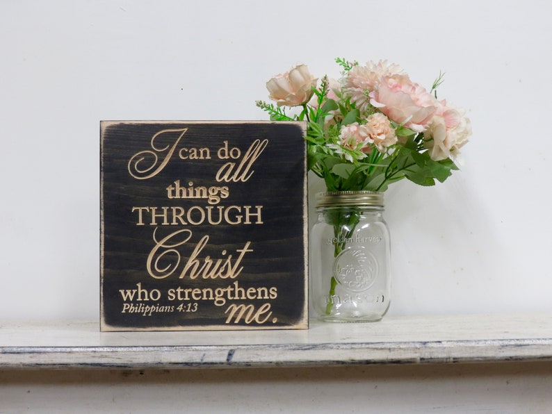 May include: A black wooden sign with the text "I can do all things THROUGH Christ who strengthens me. Philippians 4:13" in gold lettering. The sign is sitting on a white shelf with a glass jar of pink flowers.