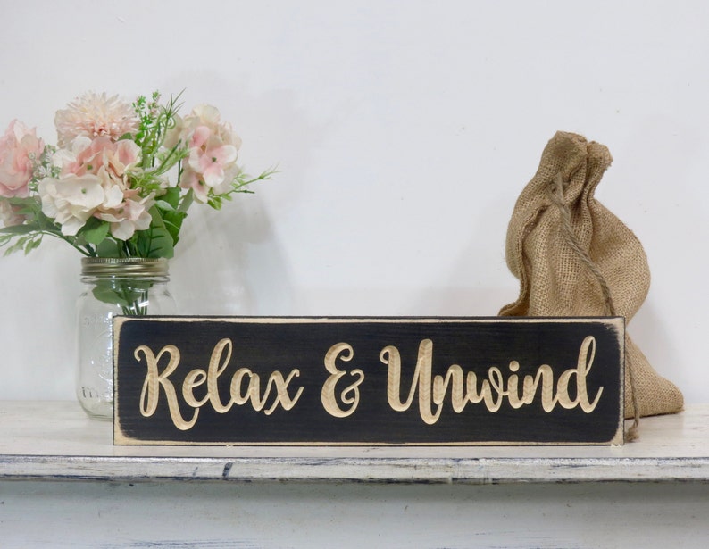 May include: Black wooden sign with white distressed edges. The sign reads "Relax & Unwind" in gold lettering.
