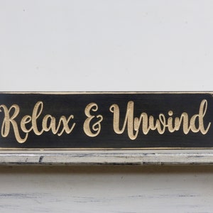 May include: Black wooden sign with white distressed edges. The sign has the words "Relax & Unwind" carved into the wood in a gold color.