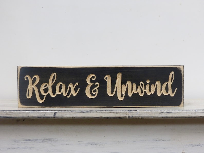 May include: Black wooden sign with white distressed edges. The sign has the words "Relax & Unwind" carved into the wood in a gold color.
