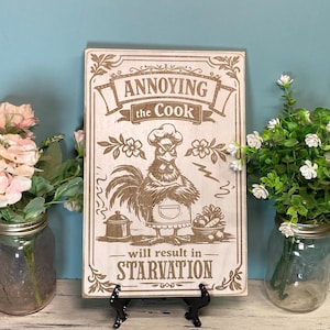 May include: Wooden sign with a rooster chef illustration and the text "Annoying the Cook will result in Starvation." The sign is brown and beige, with floral accents. The sign is displayed on a black stand, with flowers in mason jars on either side.