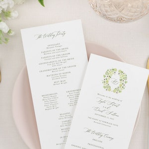 May include: Elegant wedding stationery featuring two folded programs with calligraphy text, a floral wreath, and a pink plate. Gold and white cutlery, a crystal glass, and white flowers complete the arrangement.