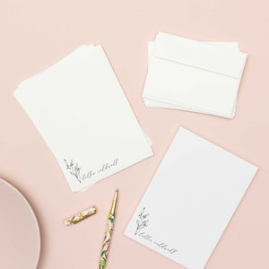 Op de afbeelding: Een set gepersonaliseerd briefpapier met briefpapier en een notitieblok. Het papier heeft een eenvoudig bloemenmotief en de naam "lilhe caldwell" gedrukt aan de onderkant. Het notitieblok heeft hetzelfde ontwerp en dezelfde naam. Het briefpapier wordt gepresenteerd op een roze oppervlak met een bloemenpen.