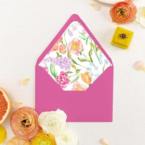 May include: A bright pink envelope with a floral patterned interior. The envelope is surrounded by flowers in shades of pink, orange, and white, along with a yellow ring box and a grapefruit slice. The floral pattern includes pink and orange flowers.