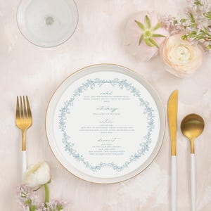 May include: A formal place setting with a white plate featuring a blue floral design, gold-handled utensils, and a menu. The setting includes a fork, knife, and spoon. Flowers and candles are also present.