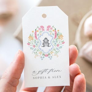 May include: A white gift tag with a floral wreath design in shades of pink, blue, and yellow. The wreath surrounds a monogram with the letters "A" and "S" in a blue and white shield. The text "a gift from Sophia & Alex" is printed below the wreath.