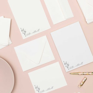 Op de afbeelding: Een platte lay-out van wit briefpapier met een bloemenontwerp en de naam "Lillie Caldwell" gedrukt op elk vel. Het briefpapier is gerangschikt op een roze oppervlak met een wit bord en een gouden pen.