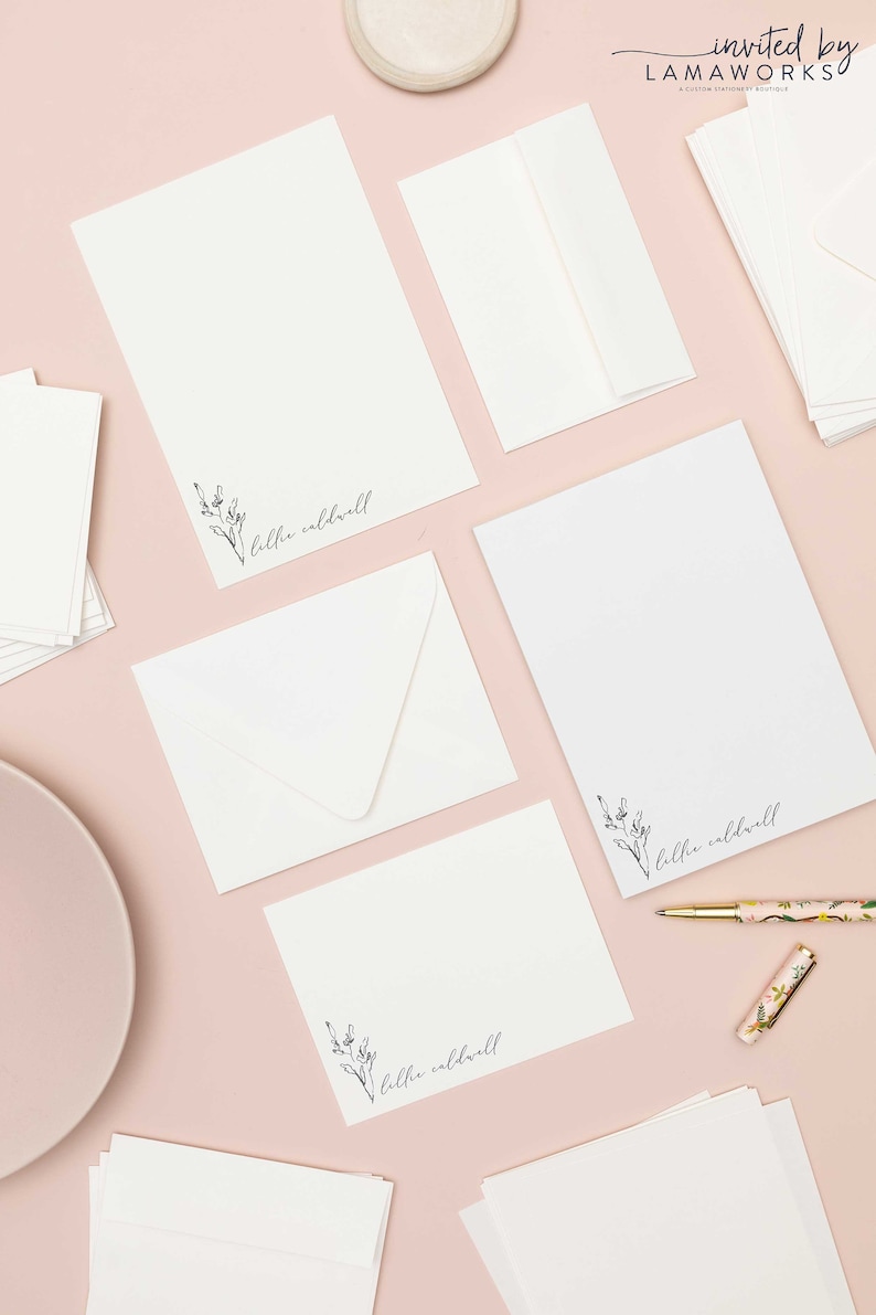 Op de afbeelding: Een platte lay-out van wit briefpapier met een bloemenontwerp en de naam "Lillie Caldwell" gedrukt op elk vel. Het briefpapier is gerangschikt op een roze oppervlak met een wit bord en een gouden pen.