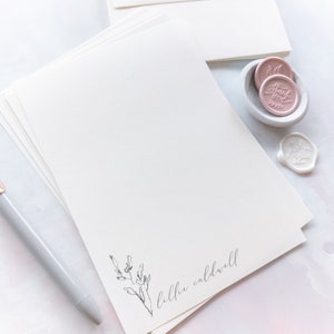 Op de afbeelding: Een stapel wit briefpapier met een eenvoudig bloemenontwerp en de naam "lilli caldwell" onderaan gedrukt. Het briefpapier wordt vergezeld door een pen en twee roze waszegels.