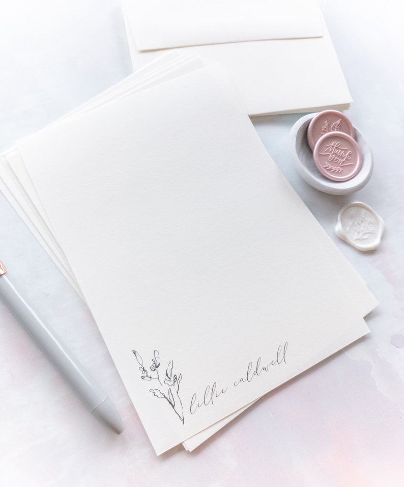 Op de afbeelding: Een stapel wit briefpapier met een eenvoudig bloemenontwerp en de naam "lilli caldwell" onderaan gedrukt. Het briefpapier wordt vergezeld door een pen en twee roze waszegels.