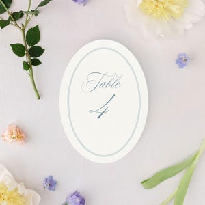 May include: An oval table number card with the word "Table" and the number "4" in a light blue script. The card is surrounded by flowers, including roses, peonies, and tulips, in shades of pink, yellow, and purple. The card is on a white surface.