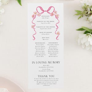 May include: A wedding ceremony program with pink ribbon and floral accents. The program lists the officiant, parents of the bride and groom, maid of honor, best man, bridesmaids, and groomsmen. It also includes a section for "In Loving Memory" and a thank you message.