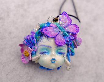 Puede incluir: Un colgante caprichoso con forma de rostro estilizado, adornado con una mariposa y detalles florales en tonos morados, azules y rosas. El colgante tiene pendientes en forma de lágrima y está suspendido de un cordón negro.