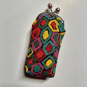 Può includere: Una custodia in tessuto rettangolare con chiusura a scatto in metallo dotata di due piccole perline rotonde color malva. L'esterno è trapuntato e decorato con un motivo geometrico ripetitivo di forme rosse, gialle, verdi e viola su fondo scuro.