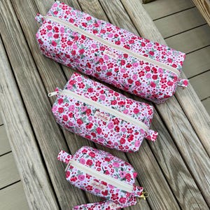 Könnte beinhalten: Drei Kosmetiktaschen mit Blumendruck und eine passende Handschlaufe. Die Taschen haben einen weißen Hintergrund mit rosa und roten Rosen und grünen Blättern. Jede Tasche hat einen beigen Reißverschluss und ein kleines Etikett mit Text.