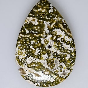 May include: A teardrop-shaped green and white gemstone with a speckled pattern. The stone is polished and has a smooth surface.
