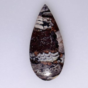 May include: A teardrop-shaped gemstone with a unique pattern. The stone features a mix of dark brown, white, and tan colors, with a textured appearance. The design resembles layers and swirls, creating a natural, organic look. The gemstone is set against a white background.